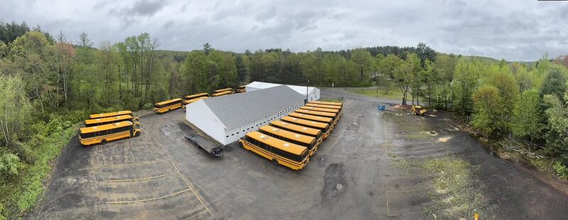 Drone photo of a school-bus depot with roughly fifteen buses parked beside a maintenance building, taken during a municipal job removing hazardous trees to keep the bus fleet in service.